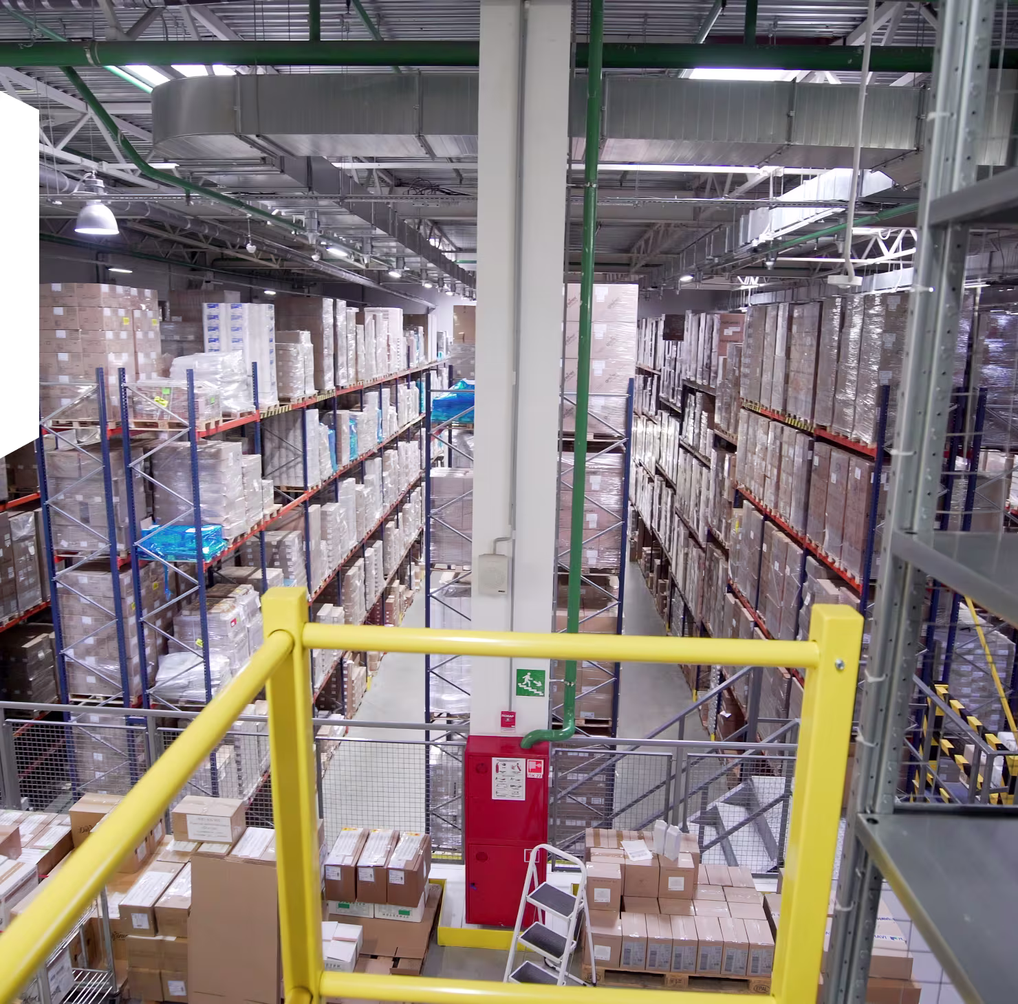 View of a spacious warehouse with tall metal shelves stacked with wrapped boxes and packages, seen from an elevated platform with yellow railings.