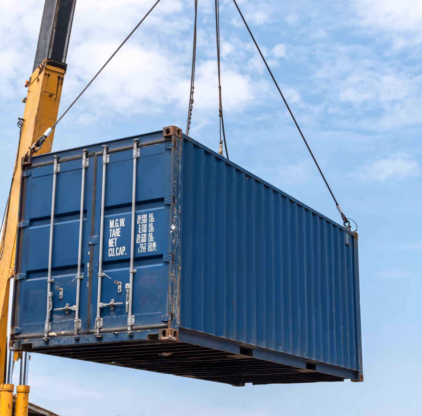 Blue shipping container being lifted by a crane against a partly cloudy sky.